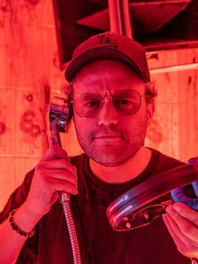 Man in cap and glasses in red-lit bathroom holding showerhead and vinyl record. Background features tiles with dark patterns.