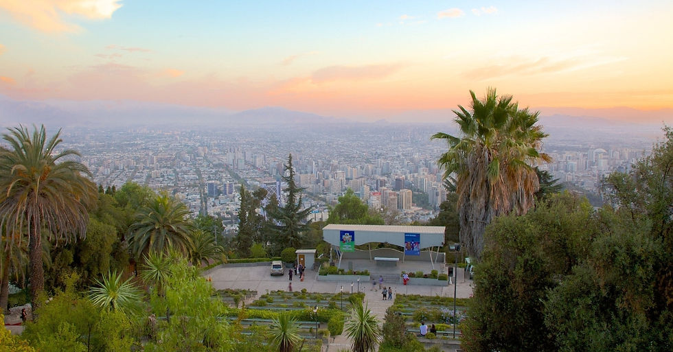 Sunset city panorama from a hilltop park with palm trees, people near a pavilion, and pastel sky over the skyline.