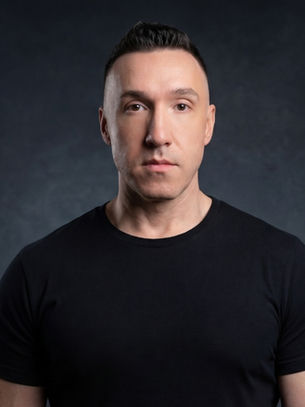 Man in a black shirt stands against a dark background with a neutral expression, creating a serious mood.