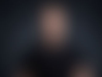 Man in a black shirt stands against a dark background with a neutral expression, creating a serious mood.