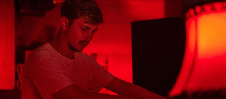 A person focused on DJing in a dimly lit, red-hued setting, with a large speaker and a lamp visible, creating an intense mood.