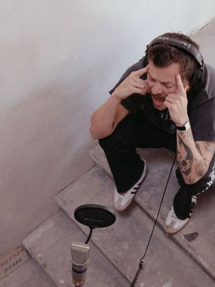 Man with headphones, sitting on steps, passionately recording into a microphone. Gray walls, small table with a phone nearby. Calm setting.