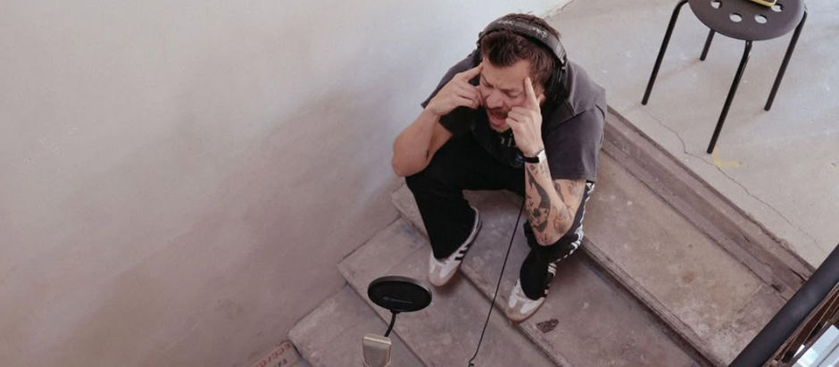 Man with headphones, sitting on steps, passionately recording into a microphone. Gray walls, small table with a phone nearby. Calm setting.