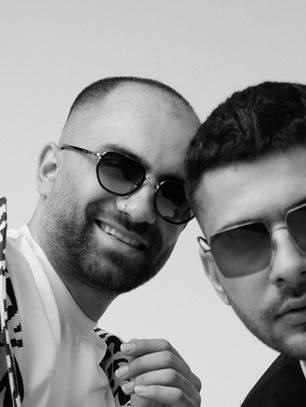 Two men wearing sunglasses pose with serious expressions. One wears a patterned jacket. Background is plain, creating a modern mood.