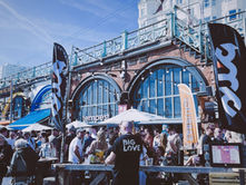 Crowd at seaside bar with arched brick facade, flags and umbrellas; man in Big Love shirt stands amid lively outdoor gathering.