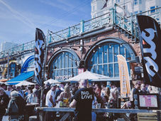 Crowd at seaside bar with arched brick facade, flags and umbrellas; man in Big Love shirt stands amid lively outdoor gathering.