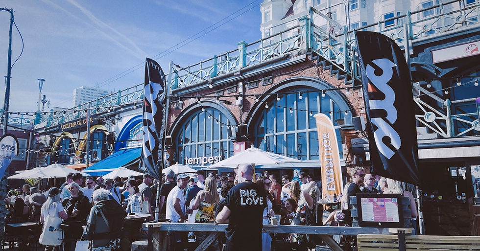 Crowd at seaside bar with arched brick facade, flags and umbrellas; man in Big Love shirt stands amid lively outdoor gathering.