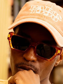 Man wearing a "PARIS 1990" cap and amber sunglasses, resting his chin on his hand in thought. Blurred background, warm tones.