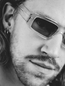 A person with long hair, wearing clear sunglasses and a nose ring, looks directly at the camera. The image is in black and white.