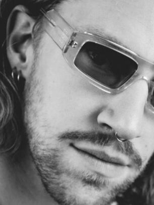 A person with long hair, wearing clear sunglasses and a nose ring, looks directly at the camera. The image is in black and white.