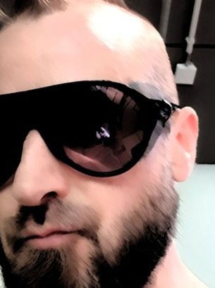 Close-up of a bearded man wearing oversized black sunglasses indoors, with an industrial ceiling behind him and a neutral expression.