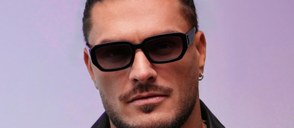 A person with dark sunglasses and a serious expression, wearing a leather jacket, against a purple gradient background.