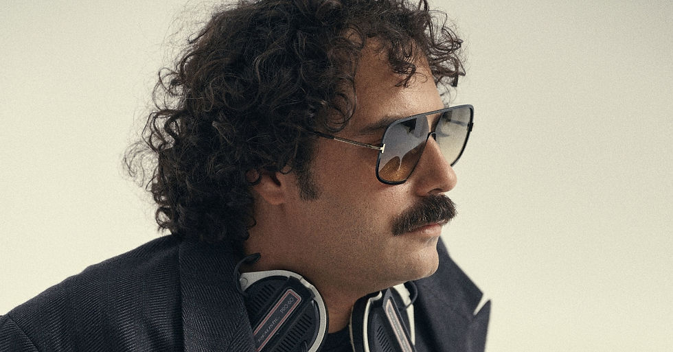 Profile of a curly-haired man with sunglasses and headphones around his neck, against a plain light background.