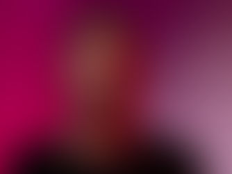 A man stands against a pink and purple gradient background, wearing a black shirt with a neutral expression. Red lighting highlights his face.