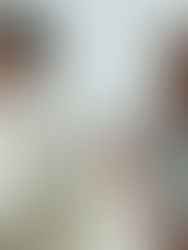 Two men in white and beige outfits sit against a plain backdrop. One adjusts his collar, the other wears a cap, both looking contemplative.