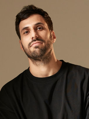 Man in a black shirt looks at the camera with a relaxed expression. Neutral beige background. Casual and calm mood.