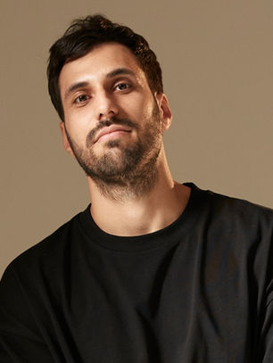Man in a black shirt looks at the camera with a relaxed expression. Neutral beige background. Casual and calm mood.