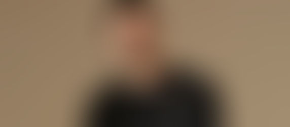 Man in a black shirt looks at the camera with a relaxed expression. Neutral beige background. Casual and calm mood.