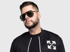 Man wearing dark sunglasses and a black jacket with white arrows on the chest, against a plain gray background, looking serious.