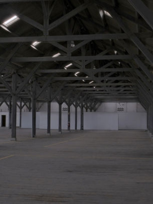 Empty industrial warehouse with wooden beams and high ceiling. Gray tone, large windows on the left, fire extinguisher visible.