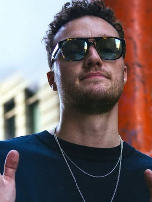 Man in sunglasses clapping outdoors, urban setting with buildings. Wearing dark shirt and chain. Confident expression, bright daylight.