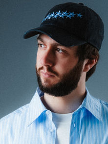 Man with a beard wearing a blue star-patterned cap and striped shirt, looking serious in a neutral, indoor setting with gray background.