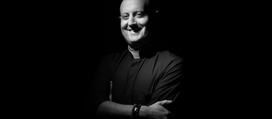 Smiling man in a priest collar stands with folded arms against a dark background. Monochrome, with focused light on his face.