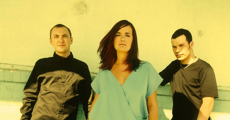 Three people pose against a pale yellow wall. The woman in blue stands between two men in black. Bright light casts strong shadows.