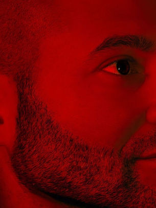 Man with a beard in a close-up profile, illuminated by red light, looking thoughtful. The background is softly blurred, enhancing the focus.