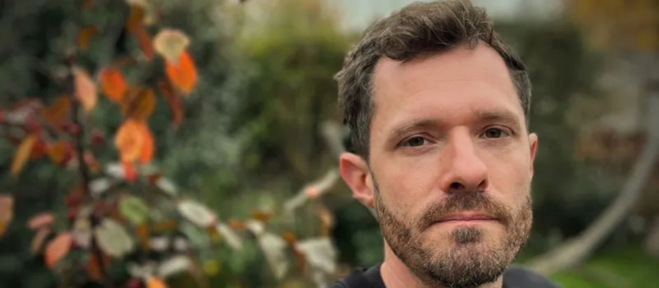 A man with a contemplative expression stands in a garden, with blurred autumn leaves in the background. Overcast sky sets a calm mood.