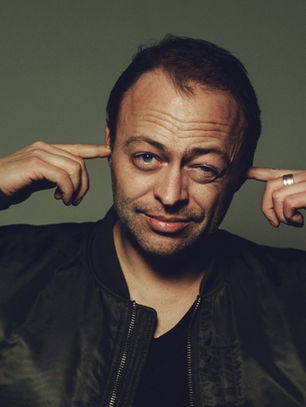 Man in a black jacket points to his ears and winks against a dark gray studio backdrop, giving a playful expression.