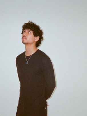 Young man in a black sweater and necklace stands against a plain wall, looking upward with a thoughtful, calm expression.