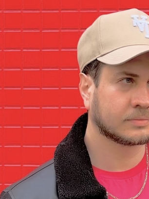 Man in beige cap and black jacket stands against red tiled wall with Japanese text, looking serious.