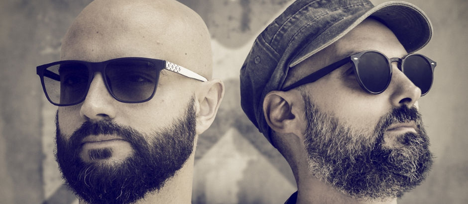 Two bearded men wearing sunglasses, one with a cap, stand against a blurred geometric background. The image has a sepia tone.