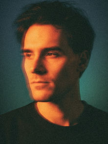 Man in a dark shirt gazes to the side under warm light. The background is a gradient of dark blue-green, creating a serene, introspective mood.