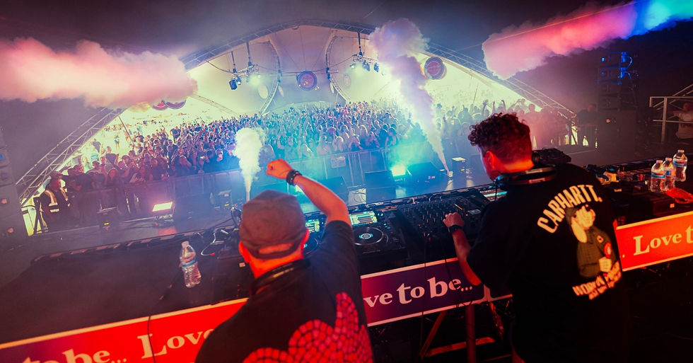 DJs performing on stage with smoke effects, vibrant lights, and a cheering crowd in a large tent. Text “Love to be…” visible on booth.