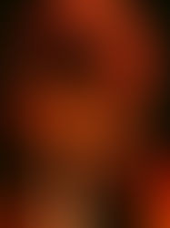Close-up of a long-haired person in a red shirt, headphones around neck, looking down under warm orange stage lighting.