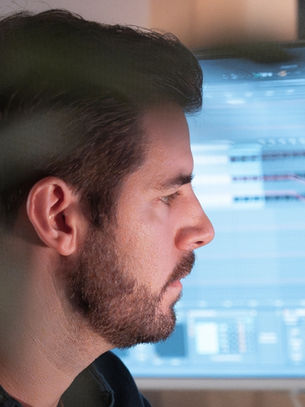 Profile of a bearded man in a dim room, focused on a blue computer monitor with audio tracks, beside a speaker and lamp.