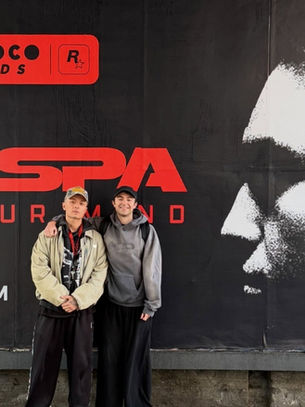 Two young men pose before a black-and-red PROSPA debut album billboard with a large monochrome face graphic.