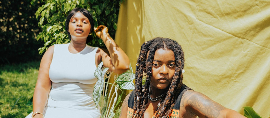 Two women pose outdoors by lush greenery and a yellow backdrop, one in a white dress, the other with dreadlocks and tattoos, serious.