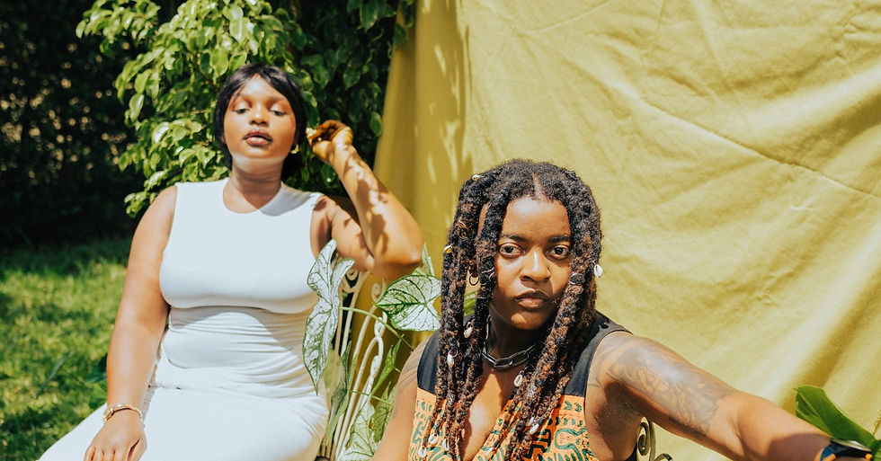 Two women pose outdoors by lush greenery and a yellow backdrop, one in a white dress, the other with dreadlocks and tattoos, serious.