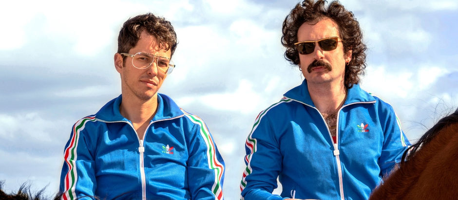 Two men in blue tracksuits and sunglasses sit on horses outdoors under a cloudy sky, looking serious.