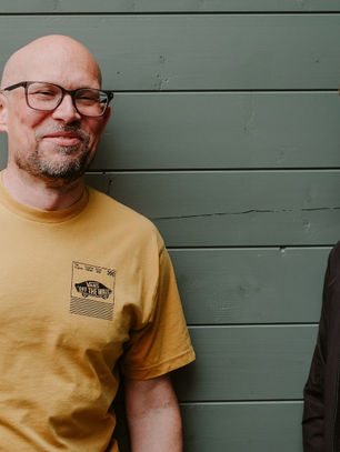 Two men stand relaxed against a green wooden wall. One wears a yellow T-shirt with text. The mood is casual and friendly.
