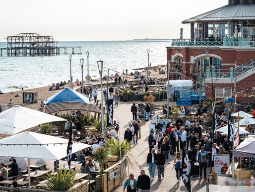 Crowd enjoying a sunny day at a seaside market with food stalls, beachgoers, and a pier in the background. Lively and bustling atmosphere.