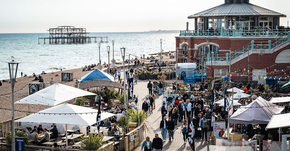 Crowd enjoying a sunny day at a seaside market with food stalls, beachgoers, and a pier in the background. Lively and bustling atmosphere.