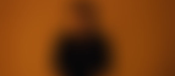 Moody studio portrait of a man in a black shirt and chain against a warm orange background, staring seriously at the camera.
