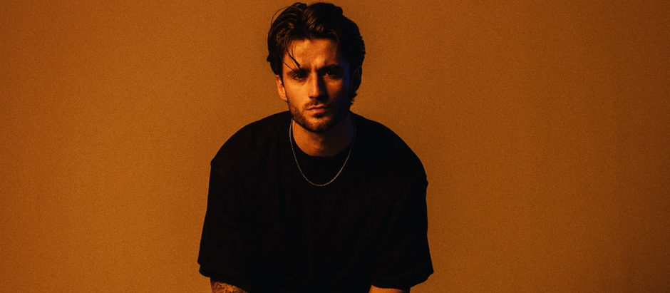 Moody studio portrait of a man in a black shirt and chain against a warm orange background, staring seriously at the camera.