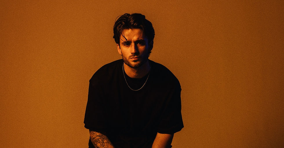 Moody studio portrait of a man in a black shirt and chain against a warm orange background, staring seriously at the camera.
