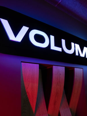 Glowing "VOLUMO" sign with bold white letters on black, set against a dark red and blue patterned background. Mood is vibrant and modern.