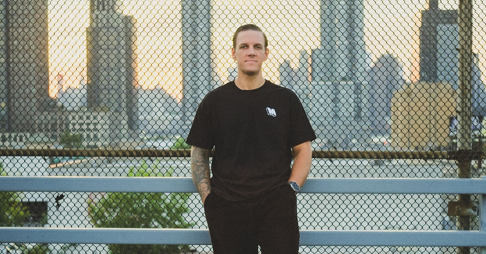 Man in black T-shirt stands by a chain-link fence on a bridge, with a city skyline at sunset behind him.